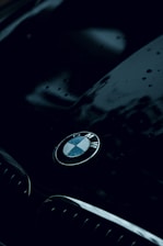 A sleek and shiny car hood is featured, with the BMW logo prominently displayed in the center. The surface appears wet with water droplets, reflecting a moody atmosphere. The dark, glossy finish of the car adds to the luxurious feel.