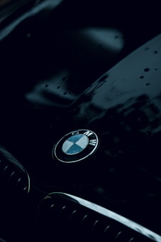 A sleek and shiny car hood is featured, with the BMW logo prominently displayed in the center. The surface appears wet with water droplets, reflecting a moody atmosphere. The dark, glossy finish of the car adds to the luxurious feel.