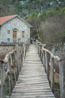 A rustic wooden bridge leads to a stone house with a red-tiled roof, set against a backdrop of dense greenery and rocky terrain. The wooden path appears worn, creating a natural, weathered look. The surrounding area is slightly overgrown, with bare branches and lush foliage creating a tranquil, secluded atmosphere.