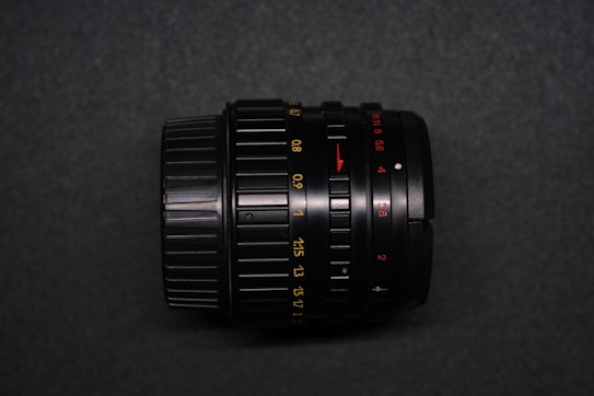 A black camera lens with various numerical markings in yellow and red on a dark surface. The lens features adjustable focus rings and textured grip areas. The background is a slightly textured gray surface, creating a contrast with the lens.