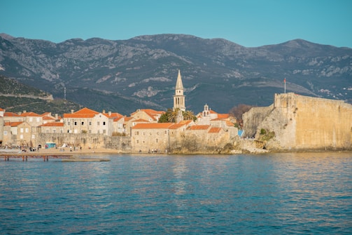 A scenic coastal town with historic stone buildings featuring red-tiled roofs. A prominent church tower rises above the structures, while a large stone wall borders the town. In the foreground, calm blue waters reflect the sunlight. Mountains form a picturesque backdrop and people are seen walking along the shoreline.