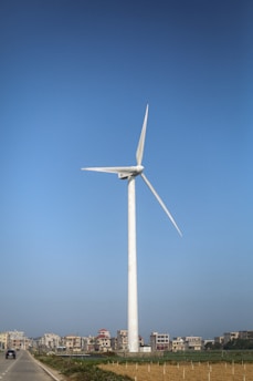 A large wind turbine stands tall in an open field, with several residential buildings visible in the background. The sky above is clear and bright blue, enhancing the sense of openness and space. To the left of the turbine, a road stretches into the distance, with a single vehicle visible, indicating a rural or less populated setting.