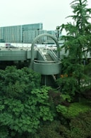 Urban scene featuring a modern architectural structure with a circular staircase surrounded by lush greenery. In the background, there are contemporary office buildings with glass facades and some vehicles parked nearby.