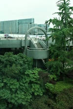 Urban scene featuring a modern architectural structure with a circular staircase surrounded by lush greenery. In the background, there are contemporary office buildings with glass facades and some vehicles parked nearby.