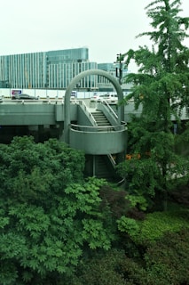 Urban scene featuring a modern architectural structure with a circular staircase surrounded by lush greenery. In the background, there are contemporary office buildings with glass facades and some vehicles parked nearby.