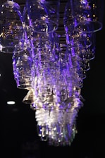 A collection of inverted glass cups hangs suspended from a rack against a dark background. The glassware is illuminated with blue and purple lighting, creating a reflective and shimmering effect.