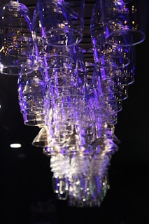 A collection of inverted glass cups hangs suspended from a rack against a dark background. The glassware is illuminated with blue and purple lighting, creating a reflective and shimmering effect.