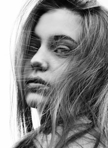 A close-up black and white portrait of a young person with long hair partially covering their face, casting soft shadows and adding texture to the image. The expression appears contemplative, with eyes gazing away from the camera.