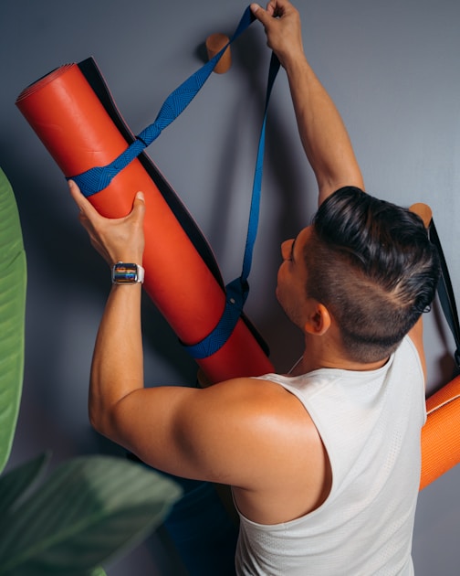 Man seen from above carrying a rolled orange yoga mat in a blue sling strap over his shoulder