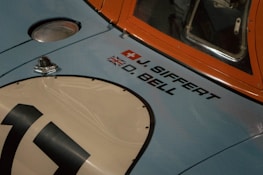 A vintage car with light blue and orange paint displays names and flags beside a racing number plate. The Swiss and Union Jack flags are above the names J. Siffert and D. Bell on the car's body.