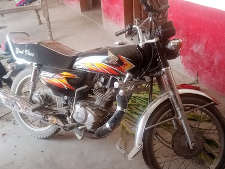 A motorcycle is parked indoors on a concrete floor, displaying custom graphics and text saying 'Good Vibes' on the seat. The motorcycle has a mix of black and orange colors with chrome details and is situated next to a red wall with some scattered leaves on the floor.