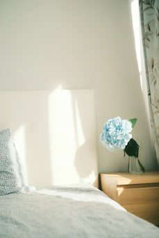 A bedroom scene featuring a neatly made bed with a patterned duvet and pillow, next to a wooden nightstand. On the nightstand, there is a clear glass vase holding a large blue hydrangea. Natural light casts soft shadows on the wall and bed, creating a serene and peaceful atmosphere.