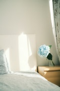 A bedroom scene featuring a neatly made bed with a patterned duvet and pillow, next to a wooden nightstand. On the nightstand, there is a clear glass vase holding a large blue hydrangea. Natural light casts soft shadows on the wall and bed, creating a serene and peaceful atmosphere.