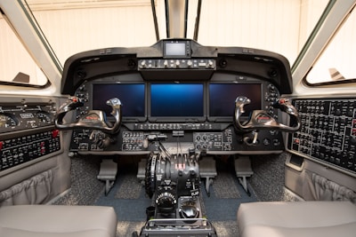 The cockpit of an aircraft featuring multiple digital displays and control panels. Two control yokes are positioned at the front, surrounded by a variety of buttons, switches, and levers. The overall design is modern with a focus on functionality and control.