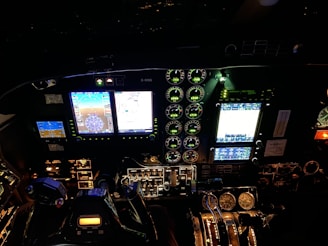 The image displays the cockpit of an aircraft at night, featuring illuminated control panels, various gauges, digital screens showing navigation and aircraft systems information, and controls such as joysticks and knobs.