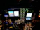 The image displays the cockpit of an aircraft at night, featuring illuminated control panels, various gauges, digital screens showing navigation and aircraft systems information, and controls such as joysticks and knobs.