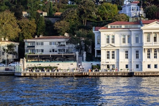 Elegant buildings with large windows are set against a lush hillside, bordering a calm waterway. A white building with classical architectural elements stands prominently next to a modern building with a patio area lined with potted plants. Several tables are arranged along the waterfront, suggesting a dining or relaxation area with a scenic view.