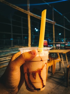 A hand is holding a plastic cup filled with a creamy beverage, likely a milkshake, adorned with chocolate syrup drizzled on the inside. A bright yellow straw protrudes from the cup. In the background, there are wooden chairs and tables, suggesting an outdoor seating area, and some string lights illuminate the area slightly, creating a cozy evening ambiance.