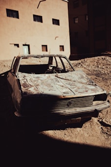 An old, rusted car with severe damage is abandoned in front of a beige building with several small windows. The area around the car appears to be a construction site or an unfinished lot, with uneven dirt ground and no visible vegetation.