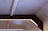 A geometric ceiling design with beams and integrated lighting. The ceiling features a rectangular frame with embedded strip lights, providing a soft, ambient glow. The materials used appear to be a combination of concrete and wood or metal, creating a modern, industrial look.