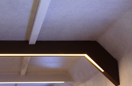 A geometric ceiling design with beams and integrated lighting. The ceiling features a rectangular frame with embedded strip lights, providing a soft, ambient glow. The materials used appear to be a combination of concrete and wood or metal, creating a modern, industrial look.