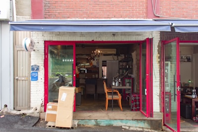 An open storefront with vibrant red framing, leading into a cozy interior with chairs and tables. The exterior features white brick walls, and various items such as boxes and bottles. A chandelier is visible inside the space.