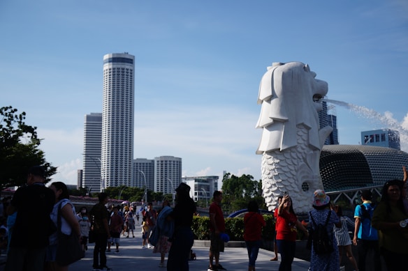 A bustling outdoor scene with many people gathered around a large, iconic statue of a lion fish hybrid, known as the Merlion. The statue is spouting water from its mouth. The background shows a cityscape with modern, tall buildings and some greenery.
