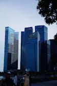 A cityscape featuring several modern skyscrapers with reflective glass facades. The buildings have logos on them, indicating corporate headquarters. People are gathered in the foreground, with trees partially framing the image.