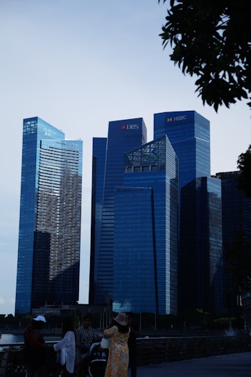 A cityscape featuring several modern skyscrapers with reflective glass facades. The buildings have logos on them, indicating corporate headquarters. People are gathered in the foreground, with trees partially framing the image.
