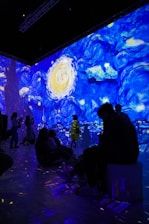 A digital art installation features projections of Van Gogh's Starry Night, casting vibrant swirls of blue, yellow, and white across the walls. People are seated and standing in the room, some silhouetted against the illuminated backdrop, creating an immersive experience.