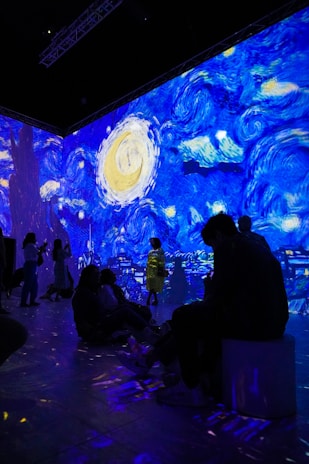 A digital art installation features projections of Van Gogh's Starry Night, casting vibrant swirls of blue, yellow, and white across the walls. People are seated and standing in the room, some silhouetted against the illuminated backdrop, creating an immersive experience.