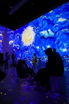 A digital art installation features projections of Van Gogh's Starry Night, casting vibrant swirls of blue, yellow, and white across the walls. People are seated and standing in the room, some silhouetted against the illuminated backdrop, creating an immersive experience.