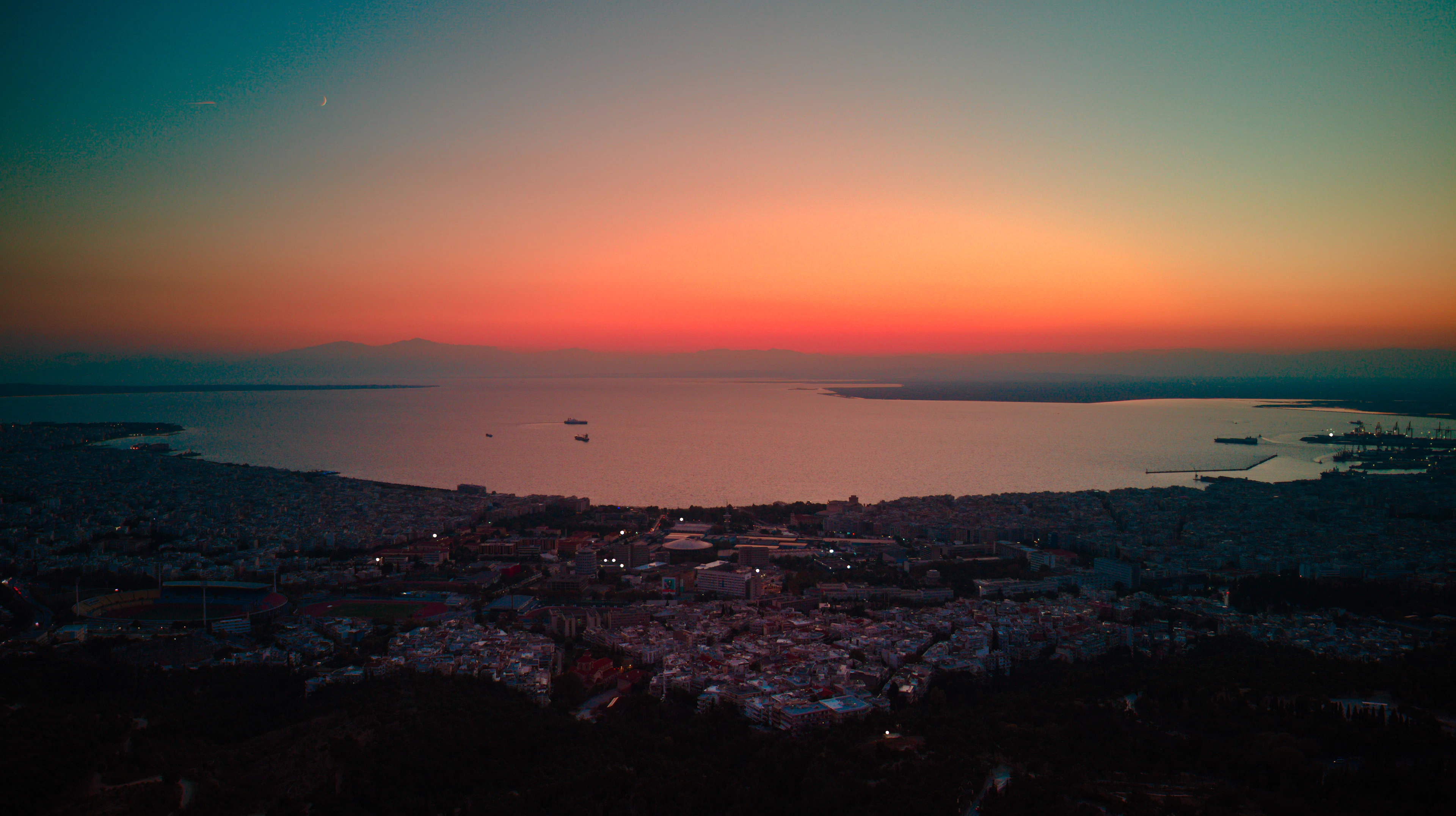 Thessaloniki at dusk from above, the lit city spread along the curve of the Thermaic Gulf under a sky graded from deep teal to burning orange, with distant mountains on the far shore.