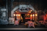 A cozy, small restaurant with a brick archway entrance is lined with red checkered tablecloths on outdoor tables. Warm, decorative lanterns hang from the entrance, casting a soft glow. Signs and bottles are visible near the entrance, suggesting a casual and inviting dining atmosphere.