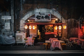 A cozy, small restaurant with a brick archway entrance is lined with red checkered tablecloths on outdoor tables. Warm, decorative lanterns hang from the entrance, casting a soft glow. Signs and bottles are visible near the entrance, suggesting a casual and inviting dining atmosphere.