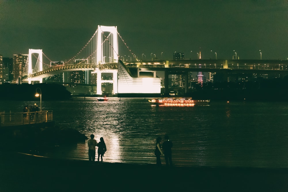 three people standing on the shore of a river in front of a bridge