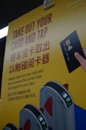 A yellow poster with instructions in English and Chinese about taking out a card and tapping it at a terminal, accompanied by an illustration of a hand holding a card and tapping it against a card reader. The poster also features illustrated turnstiles.