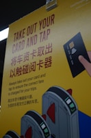 A yellow poster with instructions in English and Chinese about taking out a card and tapping it at a terminal, accompanied by an illustration of a hand holding a card and tapping it against a card reader. The poster also features illustrated turnstiles.