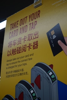 A yellow poster with instructions in English and Chinese about taking out a card and tapping it at a terminal, accompanied by an illustration of a hand holding a card and tapping it against a card reader. The poster also features illustrated turnstiles.