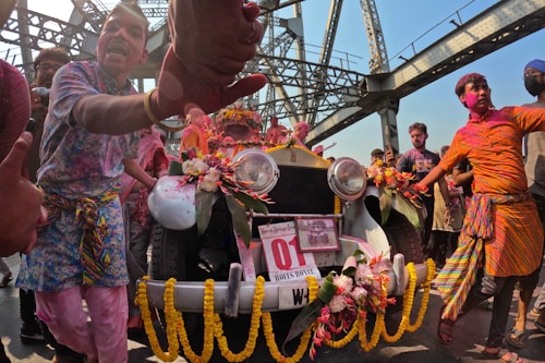A group of people are gathered around a vintage car adorned with colorful flowers and decorations, celebrating in a festive manner. The setting is outdoors beneath a large bridge. Participants are dressed vibrantly, some with colorful patterns and others with paint on their faces, suggesting a celebration or festival.
