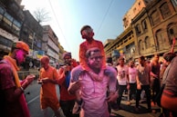 People celebrating the festival of Holi, covered in vibrant colors while walking down a bustling street lined with old buildings. A child is on the shoulders of an adult, both wearing sunglasses and smeared with colored powder. Others around are also engaging with their phones, capturing moments or conversing.