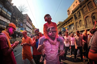 People celebrating the festival of Holi, covered in vibrant colors while walking down a bustling street lined with old buildings. A child is on the shoulders of an adult, both wearing sunglasses and smeared with colored powder. Others around are also engaging with their phones, capturing moments or conversing.