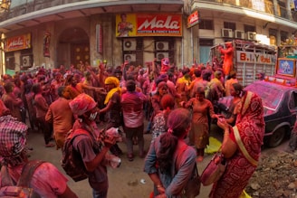 A large group of people is gathered on a street, covered in vibrant colored powders, celebrating a festival. Many participants are interacting joyfully, with some taking photos. Vibrant hues of red, pink, and yellow dominate the scene. The storefronts and a truck in the background suggest an urban setting, and traditional attire is visible on some individuals.