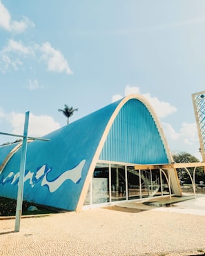 A modern architectural structure featuring a large, curved blue roof with white wave patterns. The facade includes glass doors and a series of windows. A palm tree is visible in the background, along with a clear sky with scattered clouds.