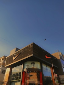 A modern building with predominantly gray and black tones featuring a large, well-known sports logo in bright orange. The facade includes glass panels displaying large advertisements. The sky is clear and blue, suggesting a sunny day. Nearby utility poles and wires are also visible.