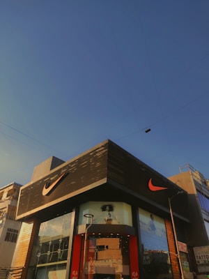 A modern building with predominantly gray and black tones featuring a large, well-known sports logo in bright orange. The facade includes glass panels displaying large advertisements. The sky is clear and blue, suggesting a sunny day. Nearby utility poles and wires are also visible.