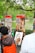 An artist is sketching a portrait in a park setting with trees overhead. The subject, an older woman, is sitting in front of the artist. Behind them are stands displaying other portraits. The scene is vibrant with greenery, and a lake is visible in the background.