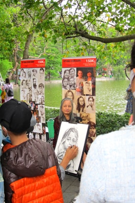 An artist is sketching a portrait in a park setting with trees overhead. The subject, an older woman, is sitting in front of the artist. Behind them are stands displaying other portraits. The scene is vibrant with greenery, and a lake is visible in the background.