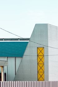 A modern industrial building featuring geometric shapes and a mix of materials. The structure has metal siding with a prominent yellow accent section in a zigzag pattern. The facade includes teal and grey panels, and there are several horizontal and vertical lines creating a minimalist architectural aesthetic.