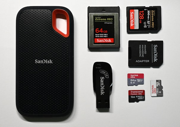 An assortment of storage devices is displayed, including a black SanDisk portable SSD with a red accent, several memory cards such as SD and microSD of varying capacities from brands like SanDisk and Transcend, along with a USB flash drive and a microSD card adapter.